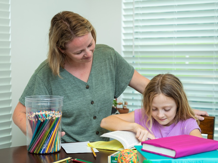 Mom helping her daughter with homework amid textbooks, colored pencils, and crayons.